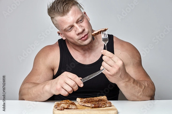 Obraz Close up big body bodybuilder eat big beef steak behind table opposite gray wall