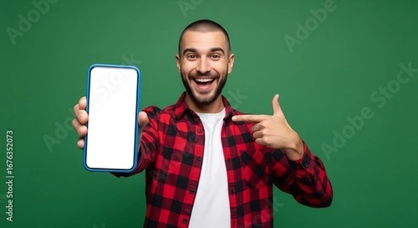 Fototapeta Happy man with beard smiling and pointing at a blank smartphone screen, wearing a plaid shirt on a green background.