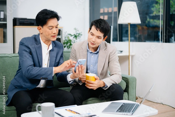 Fototapeta Two Asian businessmen relax with coffee and discuss ideas using a smartphone in a modern office.