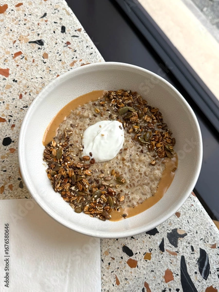 Fototapeta Oatmeal with baked granola, cream and peanut butter. Healthy breakfast and complex carbs concept. Handheld mobile photo, selective focus, non AI