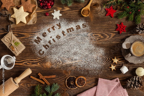 Fototapeta Wording Merry Christmas written on dark wooden background, copy space. Cookies, candle, cinnamon, cup of coffee, marshmallows, fir-tree, dried oranges. Top view.