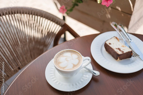 Fototapeta Cup of cappuccino on a table next to a delicious slice of cake on the terrace of a cafe, decorated with planters full of flowers.