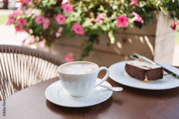 Fototapeta A white cup of cappuccino on a table next to a delicious slice of cake on the terrace of a cafe, decorated with planters full of flowers.