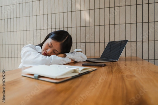 Fototapeta Young asian office worker sleeping at desk with laptop, smartphone, notebook and takeaway coffee on wooden table