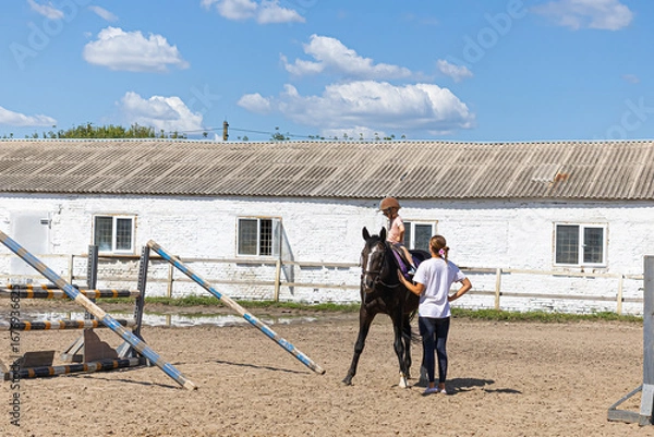 Obraz Child horse riding lesson with instructor in sunny outdoor equestrian arena