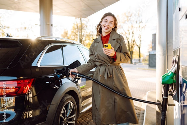Obraz A young woman with a phone fills up her car at a gas station. A woman at a gas station fills up her tank with fuel using an app on her phone. Transport concept, industry.