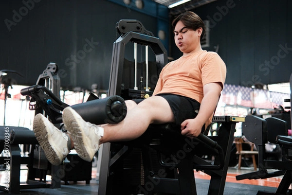 Fototapeta Overweight man exercising on a leg extension machine at the gym, focusing on lower body strength and endurance.