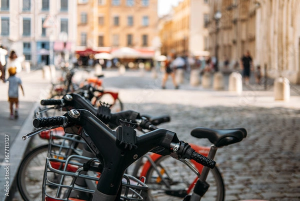 Fototapeta Row of shared bicycles parked on cobblestone street with blurred city life in the background. Urban sustainability, active transportation, car free zones, low emission commuting