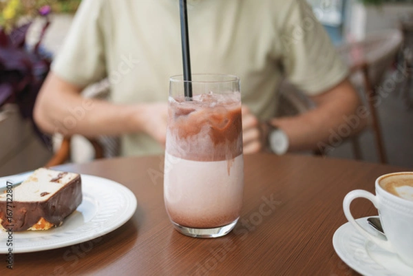 Fototapeta Close-up of a man sitting at table on summer terrace and enjoying a refreshing iced cacao drink with delicious cake.