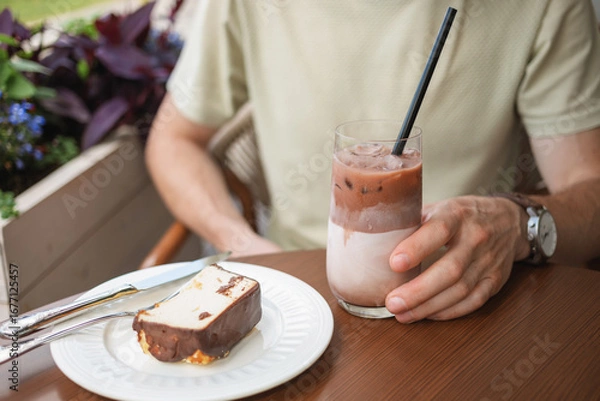 Fototapeta Close-up of a man enjoying a slice of cheesecake and a glass of iced latte coffee, while sitting on the terrace of a cafe.