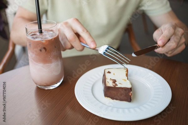 Fototapeta Close-up of a man enjoying a slice of cheesecake and a glass of iced latte coffee, while sitting on the terrace of a cafe.