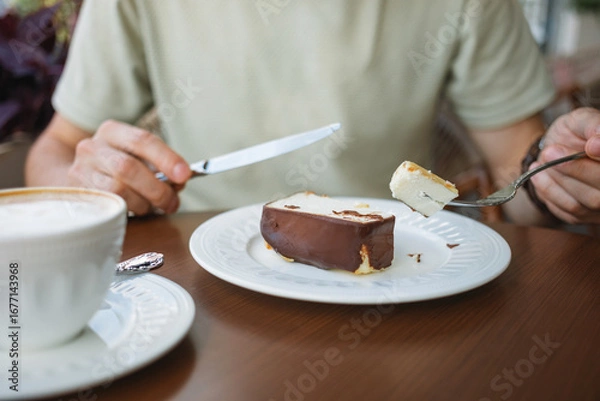 Fototapeta Close-up of a man enjoying a slice of delicious cheesecake and a cup of coffee, sitting on the terrace of a cozy cafe.
