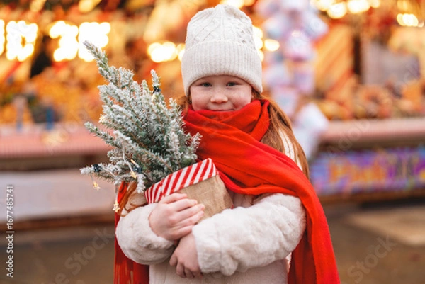 Fototapeta Child enjoys holiday festivities at a market while holding a miniature Christmas tree wrapped in red scarf and warm clothing