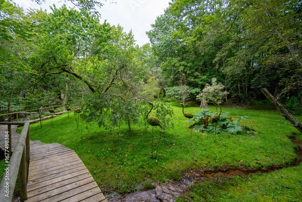 Obraz A landscaped garden park with a wooden pathway, a small stream, and decorative moss sculptures shaped like birds, surrounded by lush green forest.