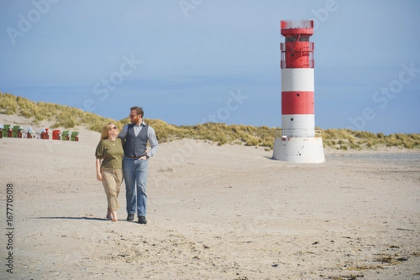 Obraz A loving couple strolls the sandy beach of Helgoland Düne with the iconic red-and-white lighthouse in the background, symbolizing romance, travel, and coastal lifestyle.