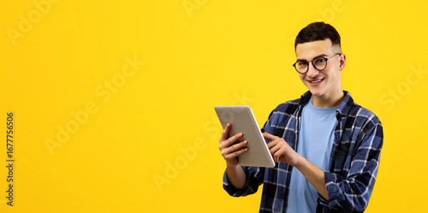 Fototapeta Online life concept. Funky young man using tablet pc on orange studio background. Joyful millennial guy with touch pad surfing web, watching videos, checking email, posting in social media