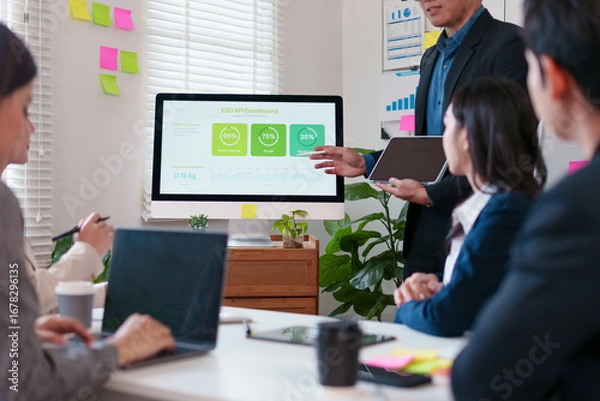 Fototapeta Team meeting in modern office with presenter pointing at ESG KPI dashboard on desktop monitor, collaborative discussion and focused colleagues engaged in strategy planning
