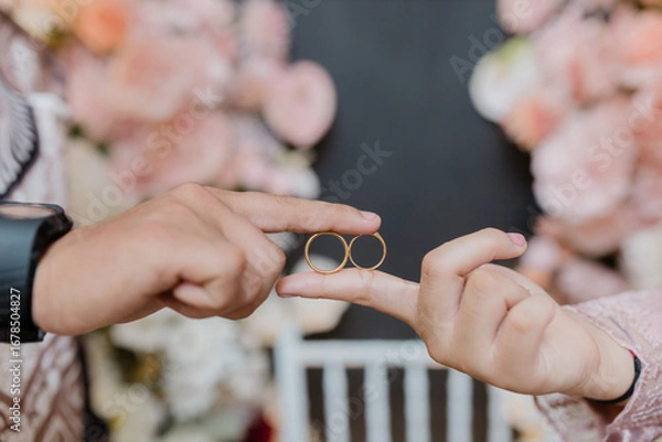 Fototapeta A close-up photo of two hands holding a pair of gold wedding rings together. One hand belongs to a man and the other to a woman, symbolizing love, unity, and commitment.