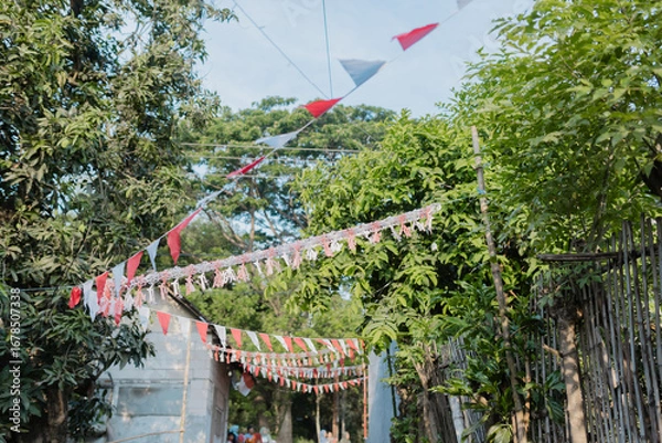 Fototapeta A festive outdoor scene in a rural neighborhood decorated with red and white triangular flags and tassels, strung across a small street. The flags, hanging between trees and bamboo fences