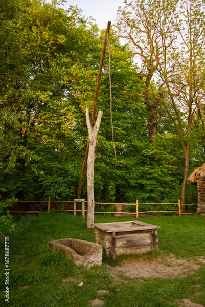 Obraz A traditional wooden water well with a stone trough, surrounded by lush green trees and grass. A great fit for historical exhibits and rustic design projects.