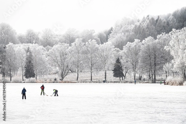 Fototapeta Laughter echoes as kids glide over the icy pond surrounded by snowy trees and fields.