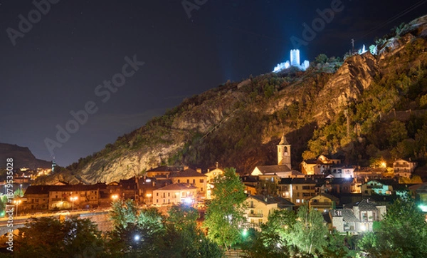 Fototapeta Night view of a town Villeneuve in the Aosta Valley in Italy. The ruins of the medieval castle Chatel-Argent, illuminated by portholes, on the top of the hill above the town. Temple and bell tower.