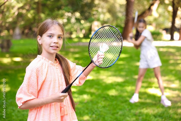 Fototapeta Smiling little girl with badminton racket and shuttlecock playing with her friend in park, selective focus