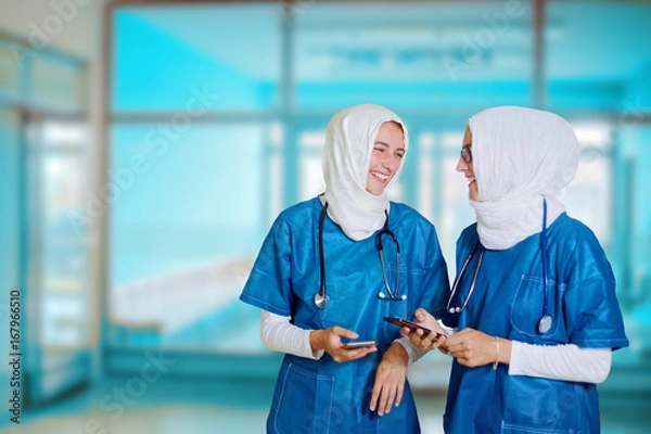 Fototapeta Two female middle eastern doctors in blue medical uniform standing in a hospital hallway, looking at each other, laughing while holding smart phones in their hands