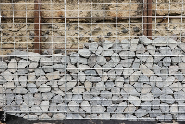 Fototapeta Stone fence in steel mesh and green lawn. Close up of gabion with tightly packed stones of varying sizes and textures. Combination of natural materials and industrial design.