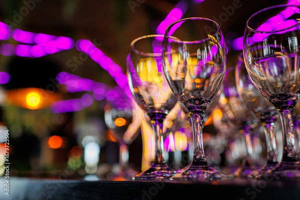 Fototapeta Empty clear wineglasses stand in rows on bar counter at nightclub, blurred purple neon and yellow lights on background. Clean cocktail glasses ready for celebrate party.