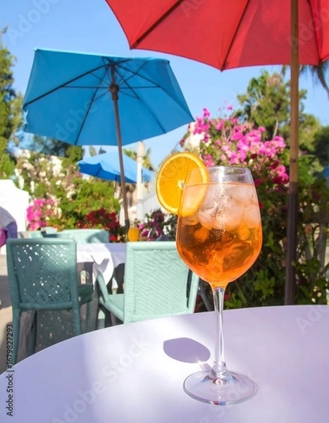 Fototapeta A vibrant orange cocktail sits on a white table under colorful umbrellas on a sunny patio.