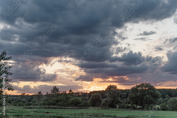 Obraz Summer evening skyscape, sun setting behind green forest and heavy dark rain clouds