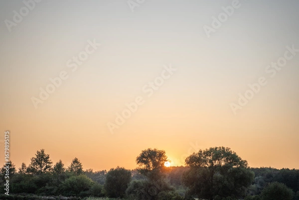 Obraz evening sky that is clear and orange at sunset, green Russian plain with forests and gardens, and setting sun