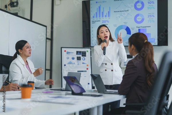 Fototapeta Businesswoman using a microphone during a presentation with charts and graphs in a meeting room with colleagues