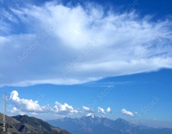 Obraz A vast, clear blue sky, dotted with soft, fluffy clouds, overlooks a mountain range, showcasing a serene and tranquil landscape.