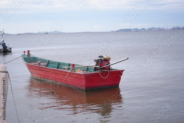 Fototapeta A traditional red long-tail wooden boat anchored by the shore, with calm sea and partly cloudy sky in the background.