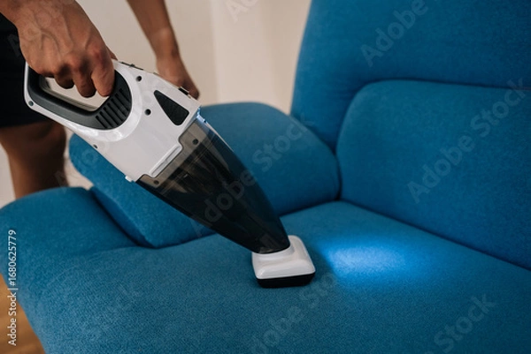 Fototapeta Close-up of cleaning service employee male using handheld vacuum cleaner to remove dust and dirt from blue sofa, emphasizing importance of cleanliness and hygiene in home maintenance.