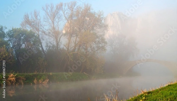 Fototapeta Misty dawn over a tranquil river, arched bridge shrouded in fog, mountains in background