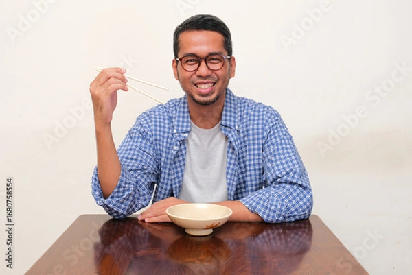 Fototapeta Adult Asian man holding chopsticks and sitting in front of empty dining bowl with happy face