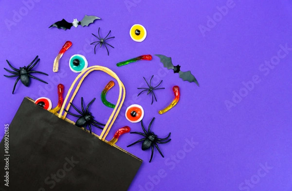 Fototapeta Black paper bag with holiday candies, gummy worms, toy spiders, bats and eyeballs on purple background. Halloween shopping and celebration concept. Top view. Copy space.