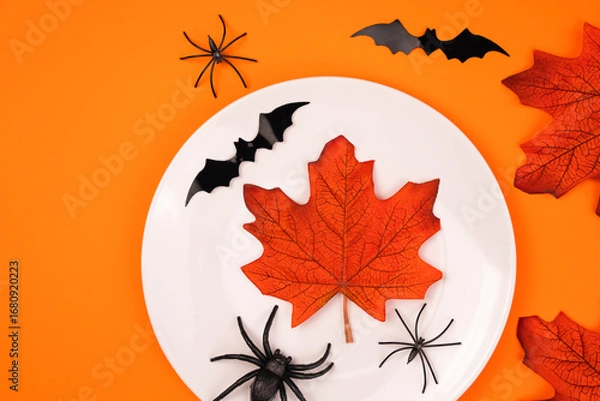 Fototapeta White plate with autumn maple leafs, spiders and black bats on bright orange background. Halloween party and table setting concept. Flatlay. Top view. Copy space.
