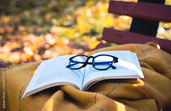 Fototapeta Open book with eyeglasses placed on a warm blanket outdoors on a bench in autumn park. Cozy fall atmosphere, reading, studying, and relaxation concept. Selective focus.