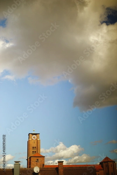 Fototapeta Red brick clock tower with a red roof and golden detail against a blue sky with dramatic white clouds. Scenic architecture view on a sunny day.