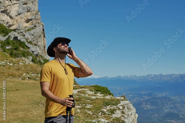 Fototapeta Male hiker in yellow t-shirt talking on phone while trekking in mountains under clear summer sky