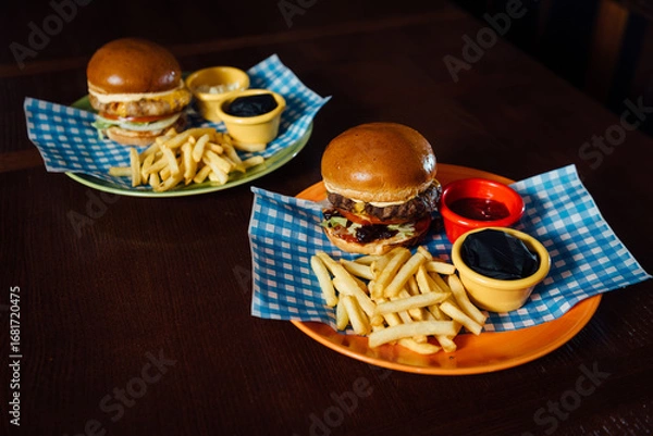 Fototapeta Two Oktoberfest burgers with tomato, cheese, and sauce, served with French fries and sauces. Presented on a plate with a Bavarian checkered napkin (lozenge pattern), evoking the spirit of Oktoberfest.