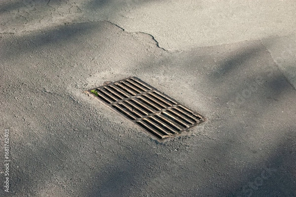 Fototapeta A storm drain grate set in old asphalt with cracks and worn textures around it.