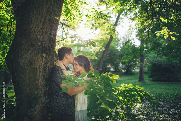 Fototapeta  Handsome young man standing in park  and kissing his girlfriend on forehead