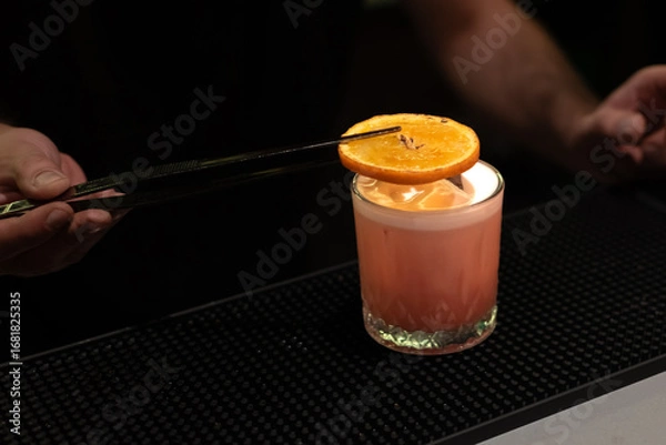 Fototapeta Bartender is using tongs to place a torched orange slice on top of a finished cocktail in a glass
