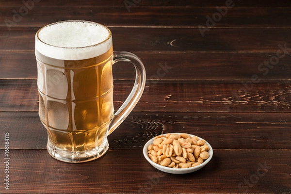 Fototapeta glass of cold frothy lager beer and peanuts plate on wooden table