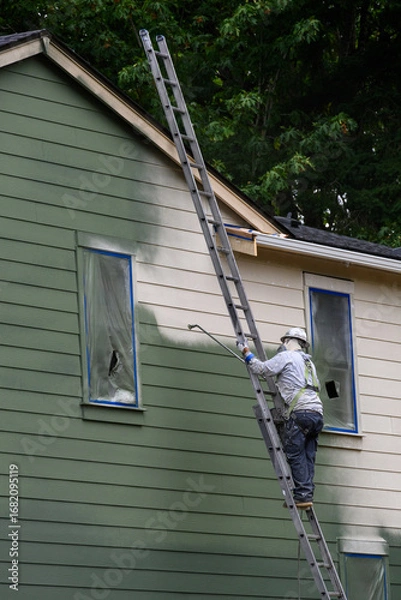 Obraz Professional painter wearing a hard hat and respirator on a ladder using a sprayer to paint the exterior of a new build house green, new housing development under construction
